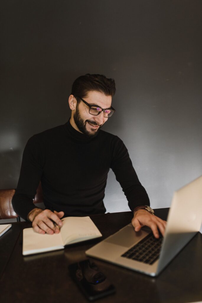 Smiling man in glasses writing in a notebook next to an open laptop, starting his first blog post