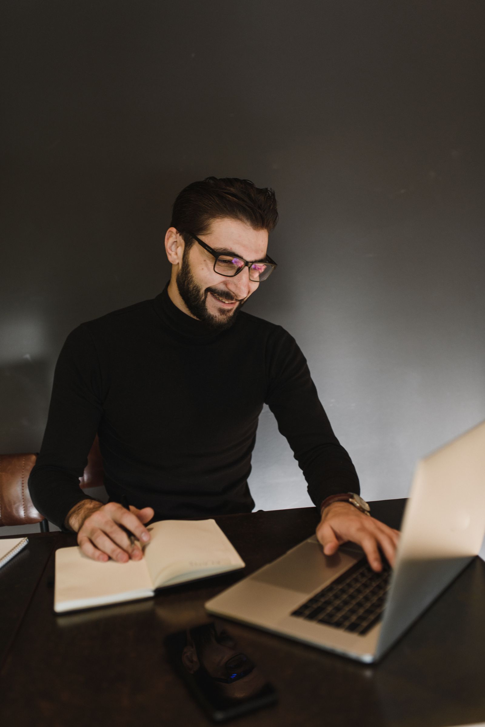 Smiling man in glasses writing in a notebook next to an open laptop, starting his first blog post