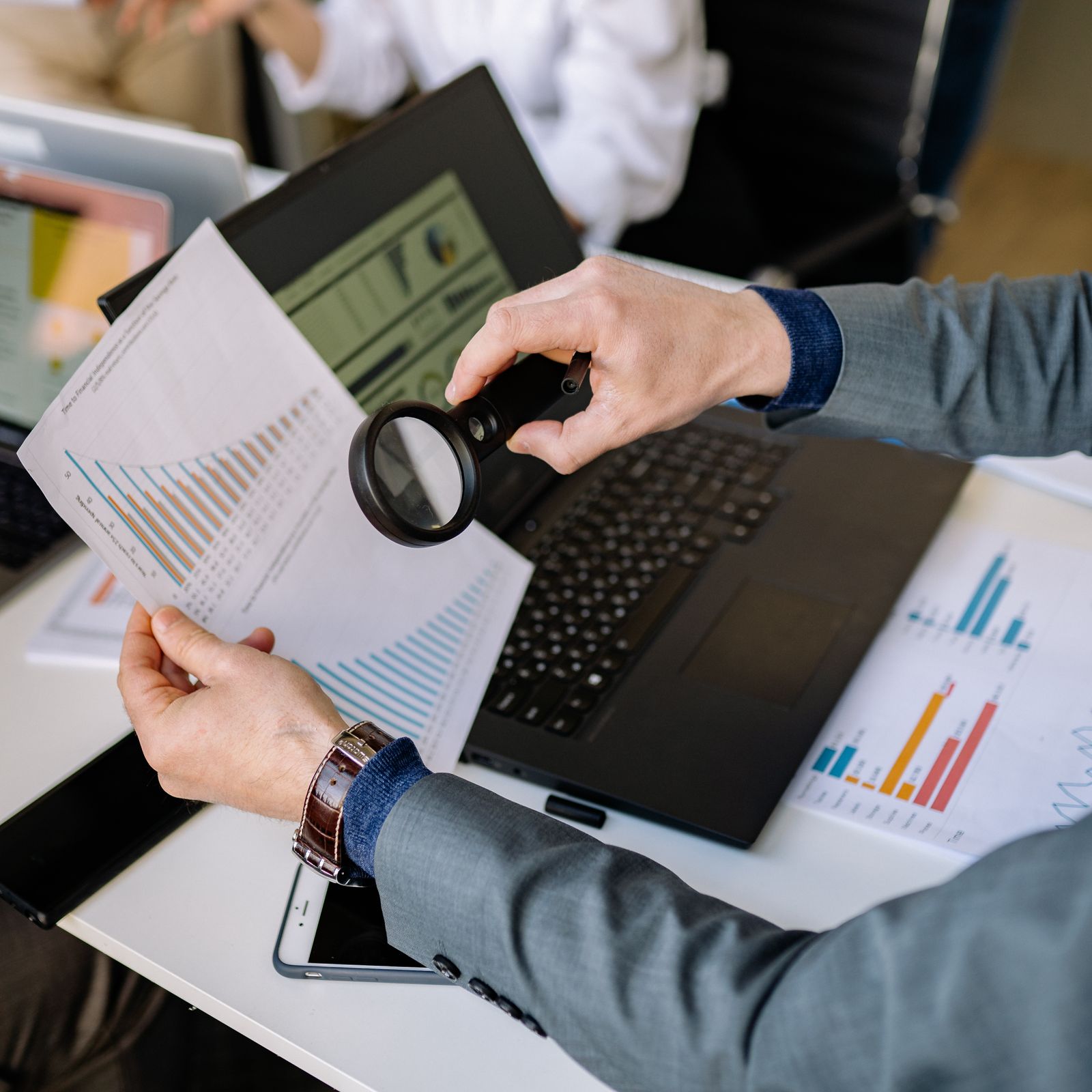 Businessman examining printed website analytics charts with a magnifying glass beside a laptop